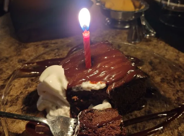 A square slice of chocolate cake, with a single red birthday candle on it.