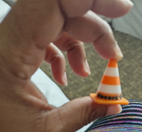 Me holding a tiny 3d printed traffic cone between thumb and forefinger. HAZARD CON is written on the bottom.