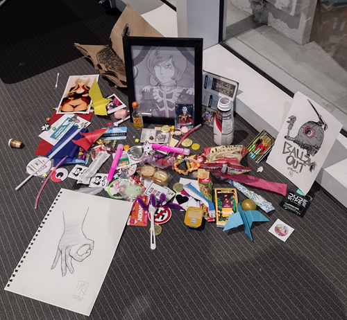 A shrine for an anime character I am not familiar with; a screenshot of his stern face in a plain black frame. People have left trinkets and libations like stickers, drawings, glowsticks, and empty Slimfast bottles.