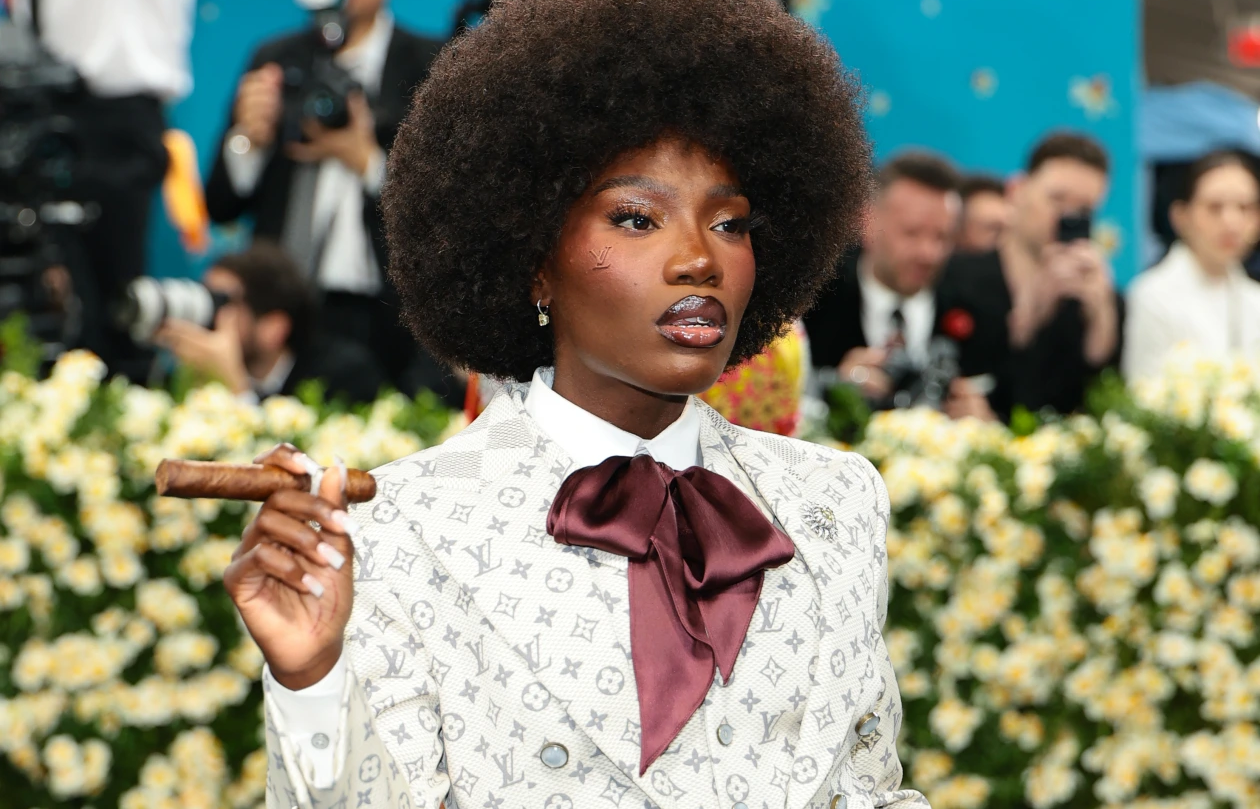 A woman with a large afro holding a cigar. Her suit is of Louis Vuitton branding, light gray on off-white, and a loose burgundy bow. Also note the scarification of the LV logo on her right cheek.