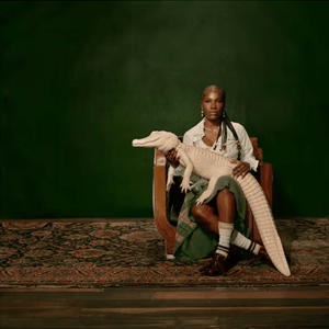 Doechii sits on a chair, facing the camera and holding an albino alligator. Behind her is a dark green wall.