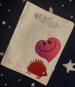 The cover of a Zine. On the top is a blue stamp of a vine. There is also a red hedgehog cutout and a pink glitterly smiling heart on the right of the cover. Title, For the Hedgie, is written on the bottom in his all-caps handwriting.